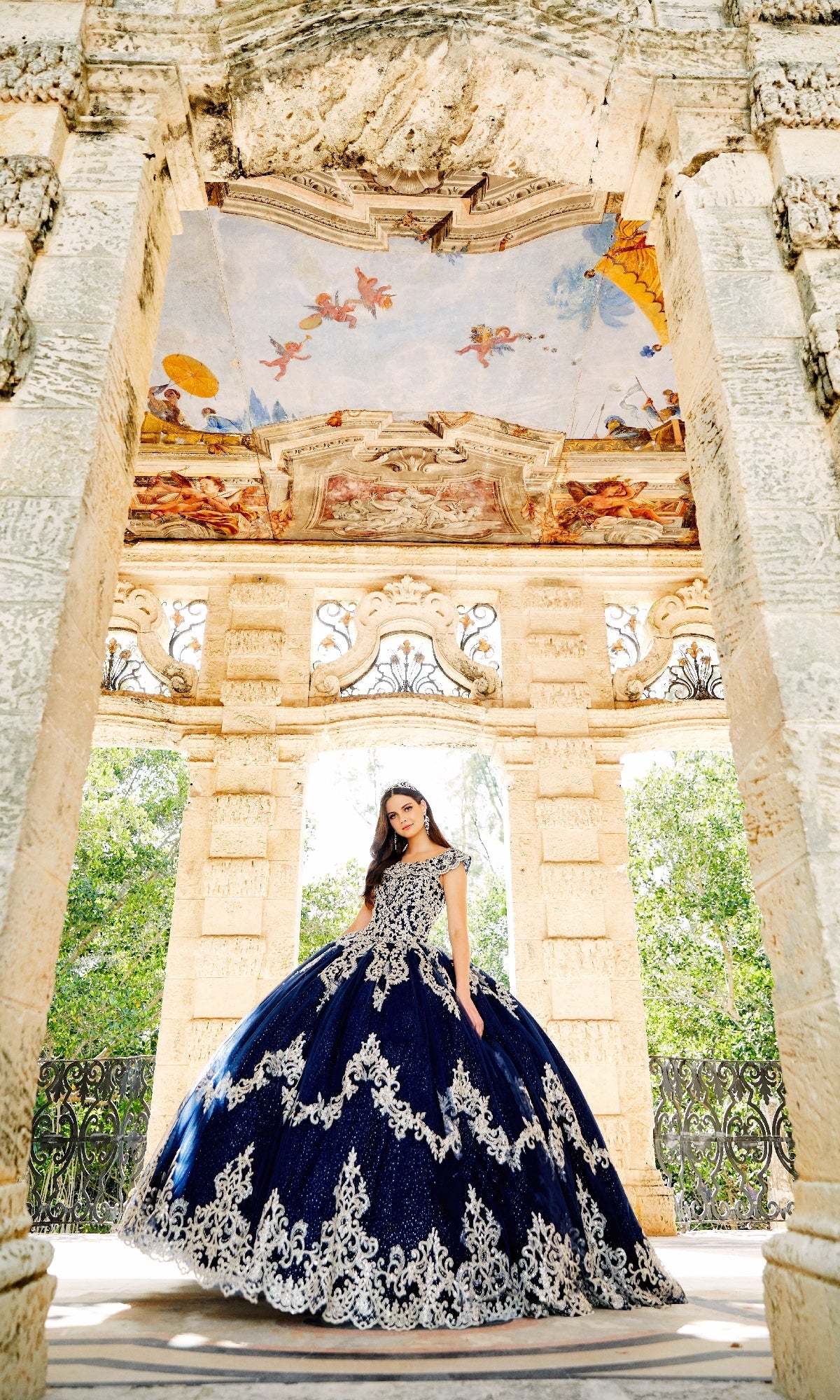 A young woman in the Quinceanera Dress PR22035 By Princesa, featuring a corset bodice and ornate navy and white design, stands in a stone gazebo with intricate ceiling artwork and lush greenery behind her.